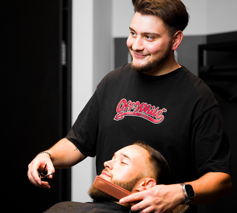 Friseur Leistungen Erkrath. Ein Mitarbeiter von Coroneo Hair Design in Erkrath ist dabei einem jungen Mann den Bart zu frisieren und zu schneiden. Sie lächeln beide. 
